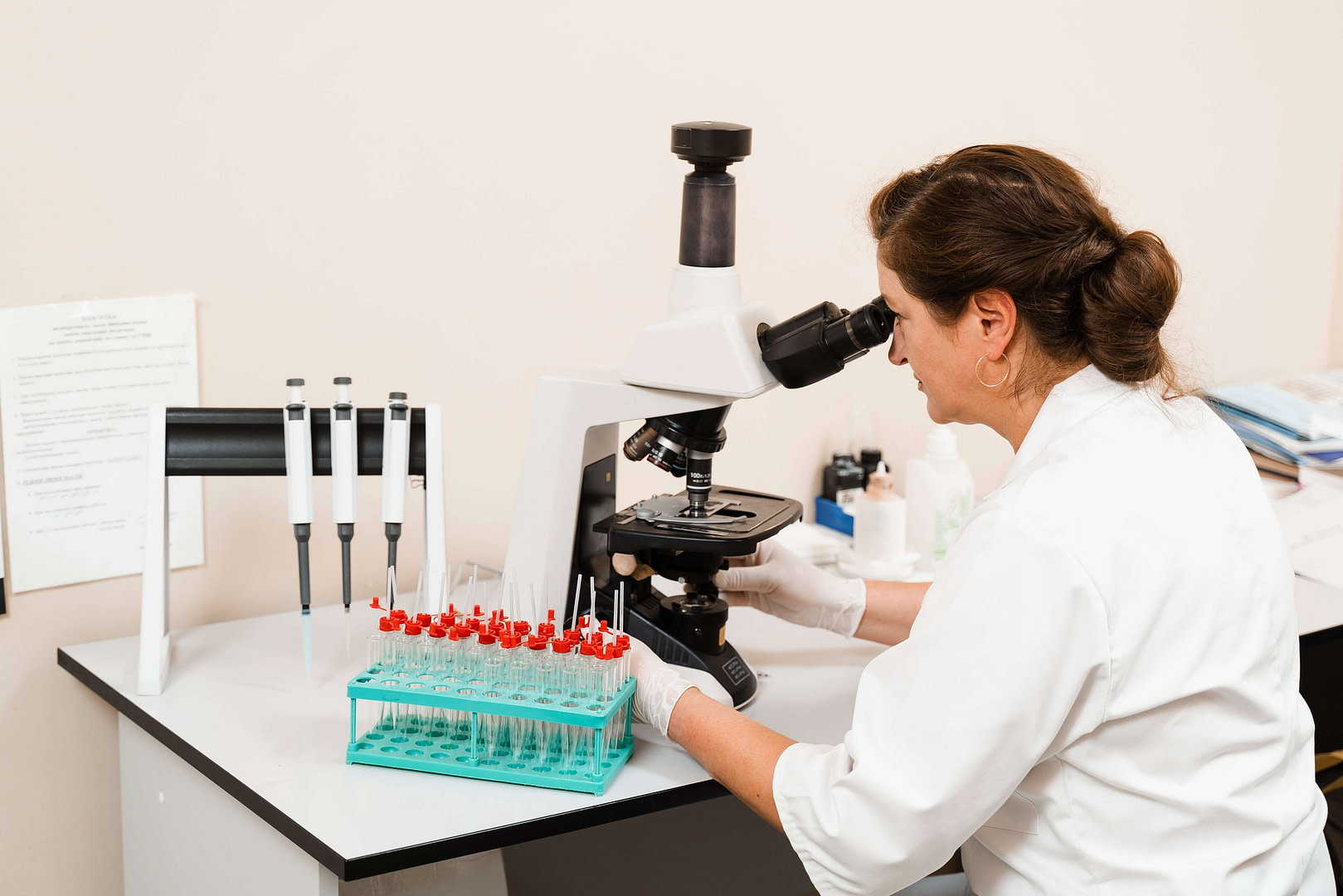 A woman in a lab coat is sat at a bench. She is looking at samples on a microscope.