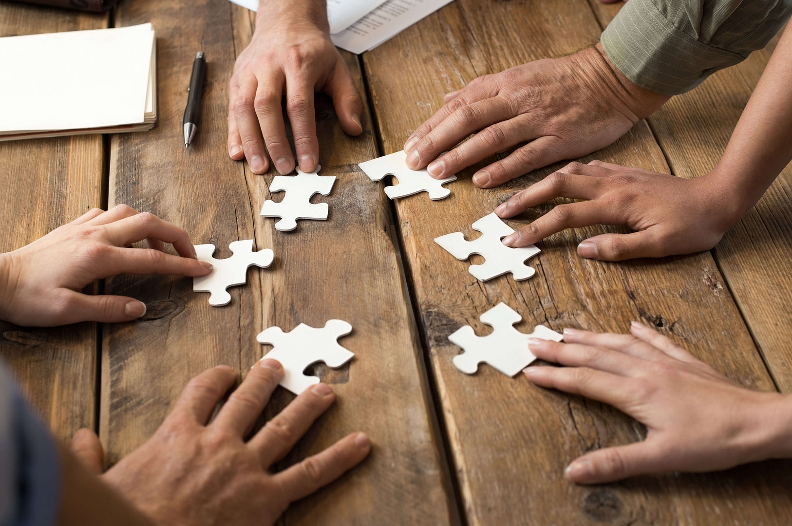 The hands of 6 different people are holding 1 piece of jigsaw puzzle each, in a circle on a table.