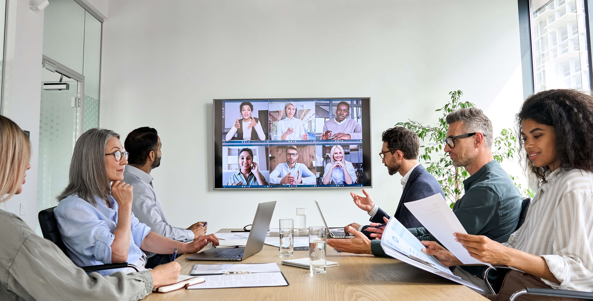 A group meeting with 12 diverse people. 6 of them are sat at a table and 6 are visible on a video conferencing screen.
